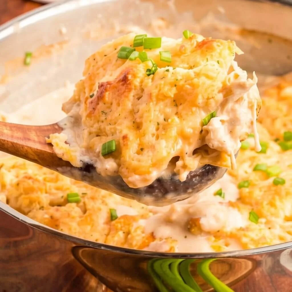 A wooden spoon scooping a portion of a creamy chicken bubble up bake from a stainless steel pan, showing chunks of chicken and gravy.