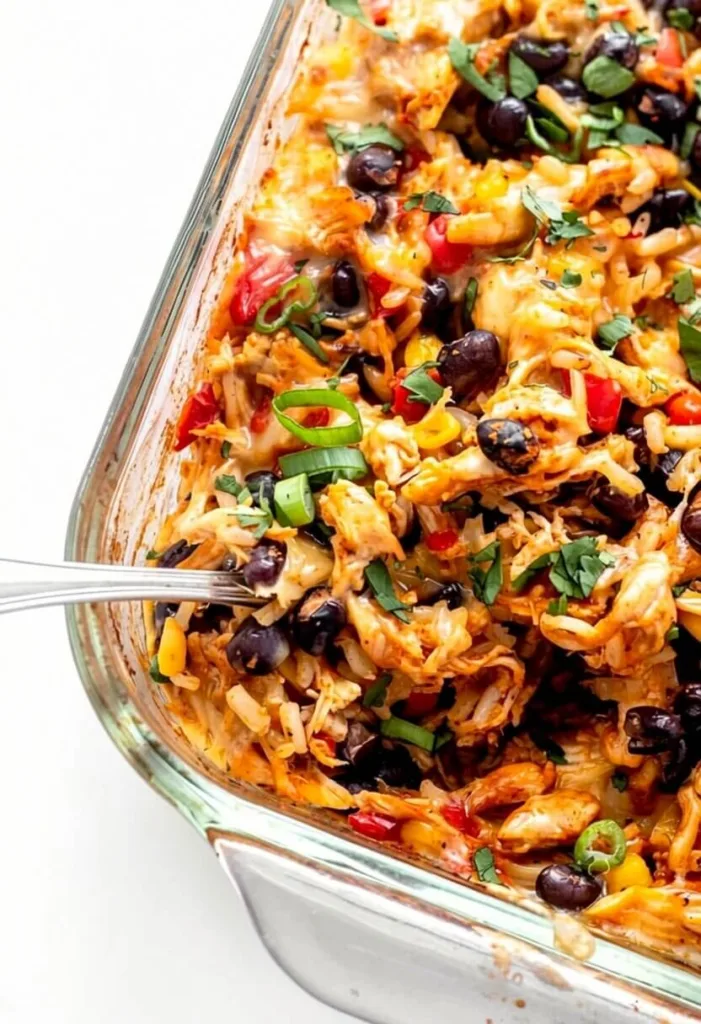 An angled close up shot of a fiesta chicken and rice bake inside a glass baking dish, being scooped with a silver spoon.